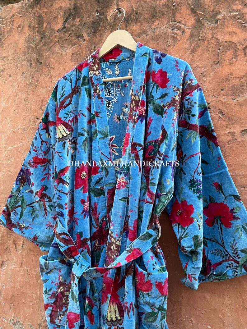 May include: A blue cotton robe with a floral print featuring red flowers, green leaves, and birds. The robe has a tie belt and long sleeves. The text "DHANLAXMI HANDICRAFTS" is printed on the robe.