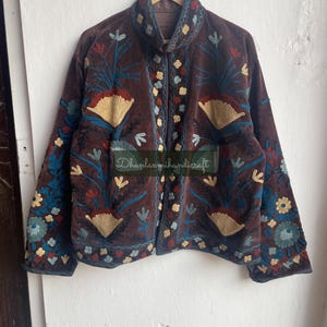 May include: A brown velvet jacket with intricate floral embroidery in shades of blue, red, and yellow. The jacket has a mandarin collar and a button closure. "Dhanlaxmihandicraft" is embroidered on the front of the jacket.
