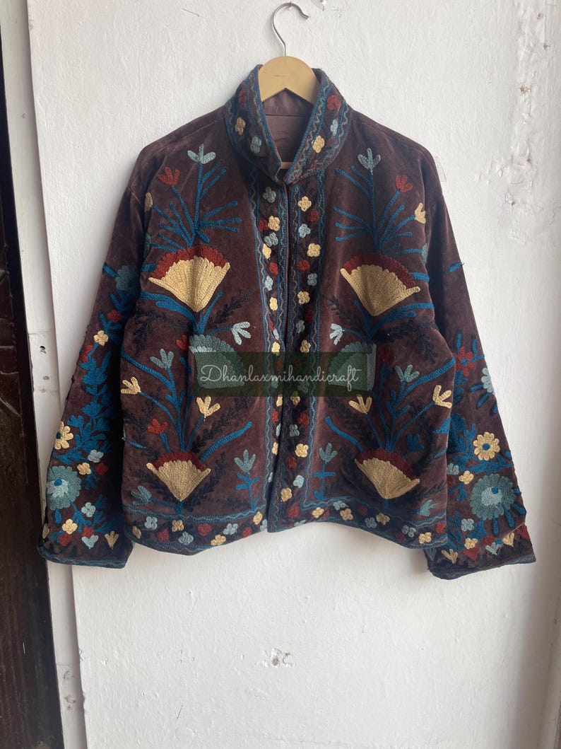 May include: A brown velvet jacket with intricate floral embroidery in shades of blue, red, and yellow. The jacket has a mandarin collar and a button closure. "Dhanlaxmihandicraft" is embroidered on the front of the jacket.