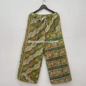 Könnte beinhalten: Weite Hose in Grün und Creme mit floralem Muster und Kordelzugbund. Die Hose hängt an einem Holzbügel. Der Text "dhanlaxmihandicraft" ist auf dem Stoff aufgedruckt. Der Hintergrund ist eine weiße Wand.