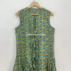 May include: Sleeveless green top with a floral pattern of yellow flowers and white leaves. The top has a ruffled hem and a tie closure at the neckline. The fabric is lightweight and appears to be cotton or a similar material.