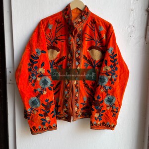May include: A bright orange velvet jacket with intricate floral embroidery in shades of blue, brown, and cream. The jacket has a mandarin collar and a button closure. The text "Dhanlaxmihandicraft" is visible on the front of the jacket.