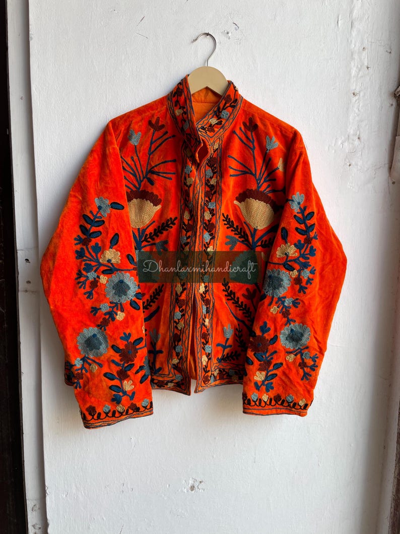 May include: A bright orange velvet jacket with intricate floral embroidery in shades of blue, brown, and cream. The jacket has a mandarin collar and a button closure. The text "Dhanlaxmihandicraft" is visible on the front of the jacket.