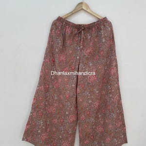 Könnte beinhalten: Weite Palazzo-Hose mit floralem Muster in Braun-, Rosa- und Weißtönen. Die Hose hängt an einem Holzbügel. Der Text "Dhanlaxmihandicra" ist sichtbar.