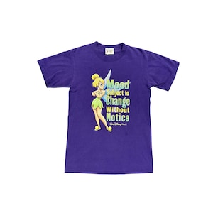 Puede incluir: Una camiseta morada con una ilustración de dibujos animados de Campanilla y la frase "Mood Subject to Change Without Notice". El diseño incluye un hada con pelo rubio y un vestido verde. La camiseta tiene cuello redondo.