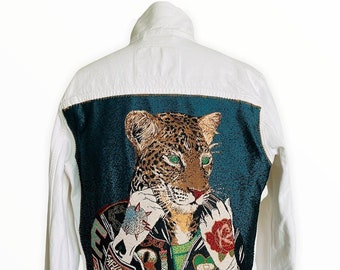 Könnte beinhalten: Weiße Jeansjacke mit einem Tapisserie-Panel auf der Rückseite, das einen Leoparden zeigt, der eine Lederjacke, ein grünes Hemd und verschiedene Tattoos trägt. Der Leopard hält eine Rose in seiner rechten Pfote.