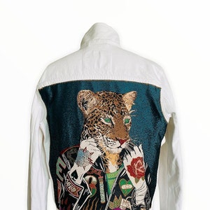 Puede incluir: Chaqueta vaquera blanca con un panel de tapicería en la espalda que representa a un leopardo con una chaqueta de cuero, una camisa verde y varios tatuajes. El leopardo sostiene una rosa en su pata derecha.