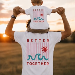 May include: White t-shirts with matching designs. The shirts feature the text "BETTER TOGETHER" in red, with pink stripes, a red sun, and teal waves. One shirt is worn by a child sitting on an adult's shoulders.
