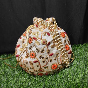 May include: A white drawstring bag with a floral embroidery design in shades of orange, red, and gold. The bag has a pearl and gold chain detail.