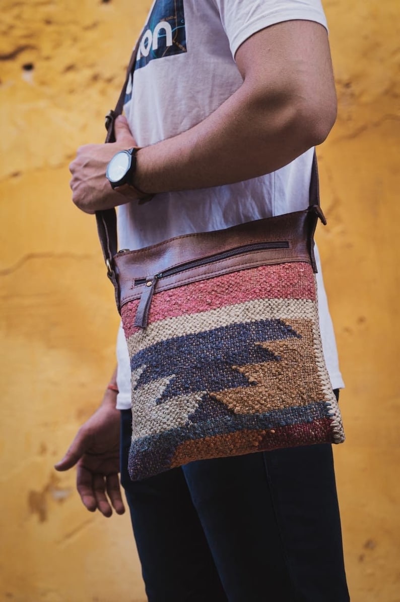 May include: A brown leather and woven crossbody bag with a zipper closure and a colorful geometric pattern. The bag is worn over the shoulder of a person wearing a white t-shirt.