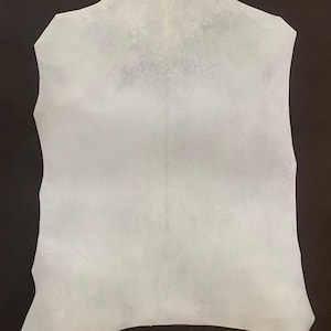 Könnte beinhalten: Ein großes, unregelmäßig geformtes Stück weißes Leder mit strukturierter Oberfläche. Das Leder wird vor einem dunkelbraunen Hintergrund präsentiert, der seine helle Farbe und natürlichen Variationen hervorhebt. Geeignet für die Herstellung.