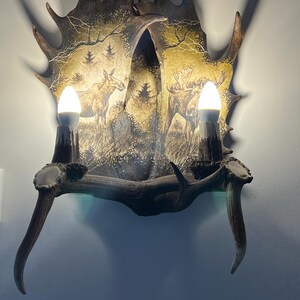 May include: A wall-mounted light fixture made from a moose antler with a painted scene of two moose in a forest. The antler is brown and has a natural texture. The light fixture has two light bulbs.
