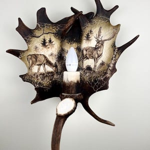 May include: A wall sconce crafted from a deer antler, featuring a light bulb and a hand-painted scene of deer in a woodland setting.