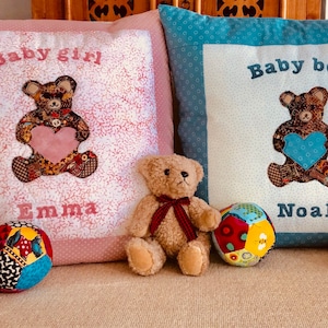 Puede incluir: Dos almohadas decorativas con osos de peluche bordados sosteniendo corazones. La almohada rosa dice "Baby girl Emma" y la almohada azul dice "Baby boy Noah". Tres osos de peluche están sentados en un sofá beige.