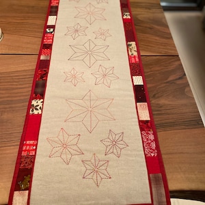 May include: A white table runner with a red border and embroidered red stars. The border is made of patchwork fabric with various patterns and colors, including red, white, and brown. The text "MERRY CHR" and "LA LA LA" is visible on the border.