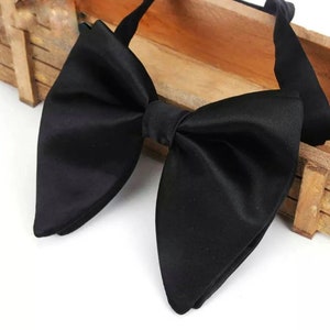 Può includere: Un papillon nero in raso, dalla classica forma a farfalla. Il papillon è realizzato in un tessuto lucido e liscio ed è esposto su una superficie di legno. È un accessorio formale.