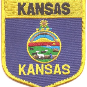 May include: Embroidered patch featuring the word "KANSAS" in yellow and black, above and below a shield design. The shield has a blue background with the state seal in the center, surrounded by a gold border.