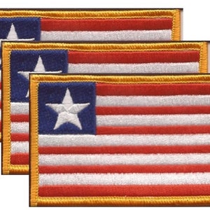 May include: A stack of embroidered patches featuring a flag design. Each patch has a blue square with a white star in the upper left corner, red and white stripes, and a gold border. The patches are rectangular.