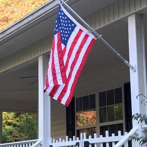 May include: An American flag with red and white stripes and a blue field of white stars is mounted on a silver pole. The flag is displayed on a white porch with a white railing and black shutters.