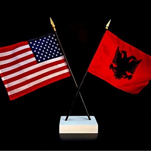 May include: Two small flags crossed at the base. The flag on the left is the American flag, with red and white stripes and a blue field of white stars. The flag on the right is red with a black double-headed eagle. The flags are on black poles.