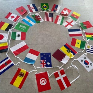 May include: A circular arrangement of fabric pennant flags representing various countries. The flags are attached to a white string, with each flag displaying the national colors and emblems. The flags are laid out on a light-colored surface.
