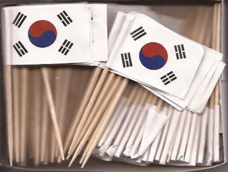 May include: A box filled with small flags of South Korea. Each flag features the Taegeuk symbol in the center, with a red and blue circle, and four black trigrams. The flags are attached to wooden toothpicks.