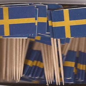 Puede incluir: Una caja llena de banderas suecas en palillos de dientes. Las banderas tienen un fondo azul con una cruz amarilla. Los palillos son de madera.