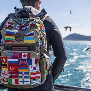 May include: A tan backpack covered in patches of various national flags. The backpack is worn by a person standing on a boat, with a view of the ocean and birds in the background. The backpack has multiple compartments and straps.