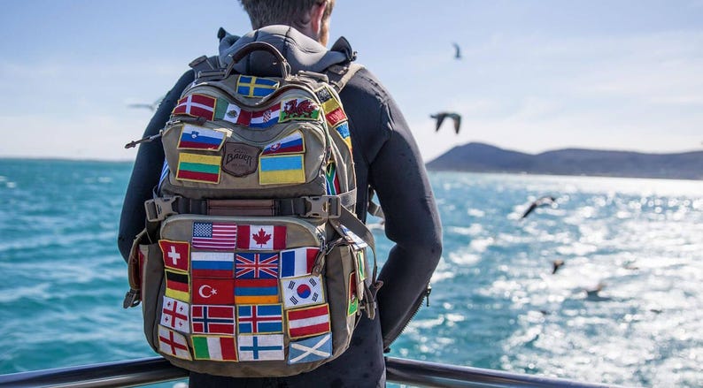 May include: A tan backpack covered in patches of various national flags. The backpack is worn by a person standing on a boat, with a view of the ocean and birds in the background. The backpack has multiple compartments and straps.