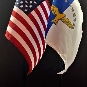 May include: Two small flags on stands. The flag on the left is the American flag with red and white stripes and white stars on a blue background. The flag on the right has a blue background with a yellow bird and stars.