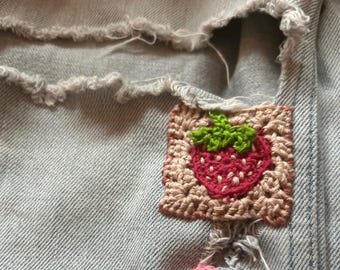 May include: A pair of light blue denim jeans with a crocheted strawberry and heart patch. The strawberry is red with green leaves and the heart is pink.
