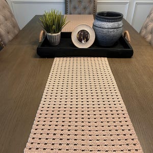 Può includere: Un runner da tavolo in crochet rosa chiaro con un vassoio nero su un tavolo in legno. Il vassoio contiene una piccola pianta in vaso, una cornice rotonda e un vaso in ceramica grigio.