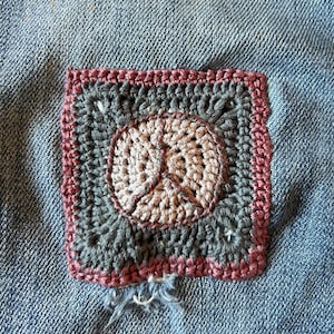 May include: A crocheted square patch with a brown, pink, and gray design. The patch is sewn onto a blue denim fabric.