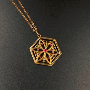 Könnte beinhalten: Goldfarbene Halskette mit Anhänger im geometrischen Design. Der sechseckige Anhänger zeigt ein Blumenmuster mit kleinen roten Edelsteinen. Die Kette ist filigran und goldfarben.