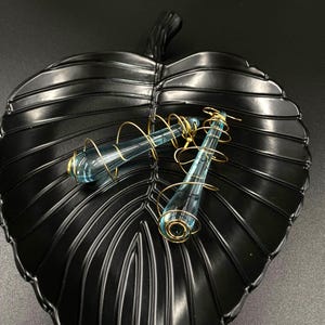 Könnte beinhalten: Ein Paar blaue Glasohrringe mit goldenen Drahtdetails, die auf einer schwarzen, blattförmigen Schale liegen. Die Ohrringe haben eine Tropfenform mit goldenen Akzenten an der Ober- und Unterseite und sind mit feinem Golddraht umwickelt.