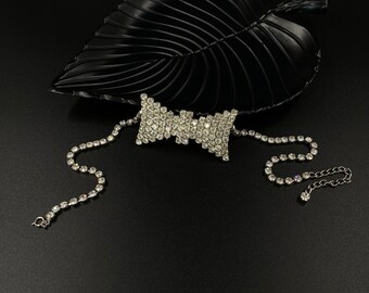 May include: A silver choker necklace with a large, sparkling rhinestone bow. The necklace is on a black background.