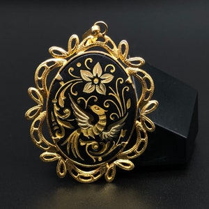 May include: A gold-tone pendant with a black oval cameo featuring a stylized bird and floral design. The cameo is surrounded by a delicate gold-tone filigree frame.