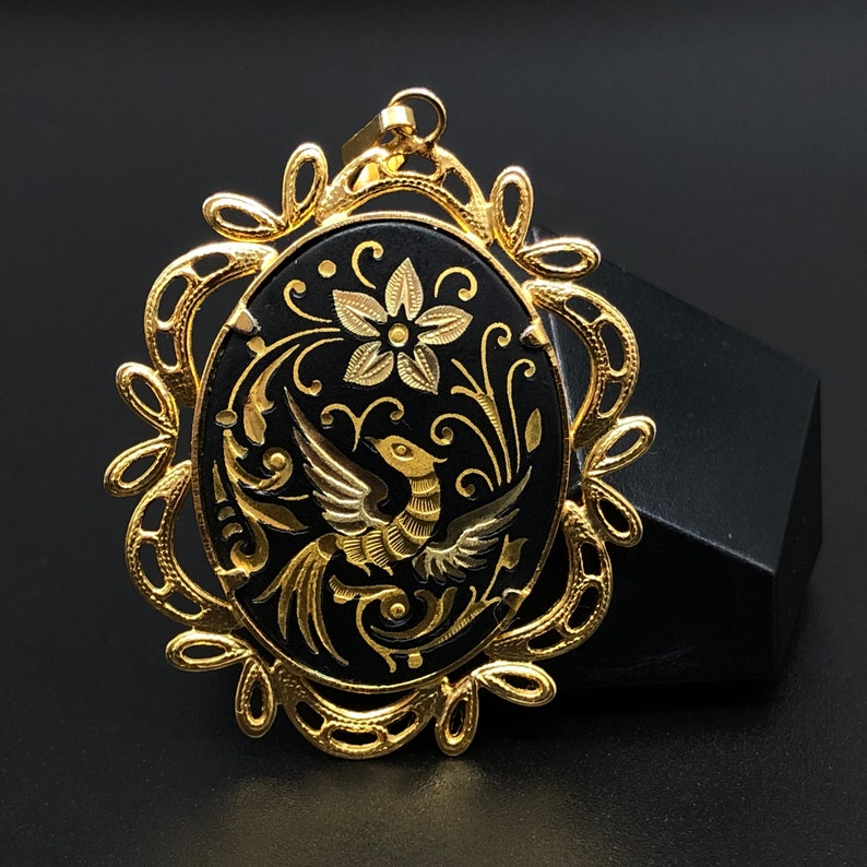 May include: A gold-tone pendant with a black oval cameo featuring a stylized bird and floral design. The cameo is surrounded by a delicate gold-tone filigree frame.