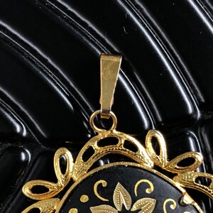 May include: A gold pendant with a black enamel center featuring a floral design. The pendant has a detailed, ornate frame and a loop for attaching a chain.