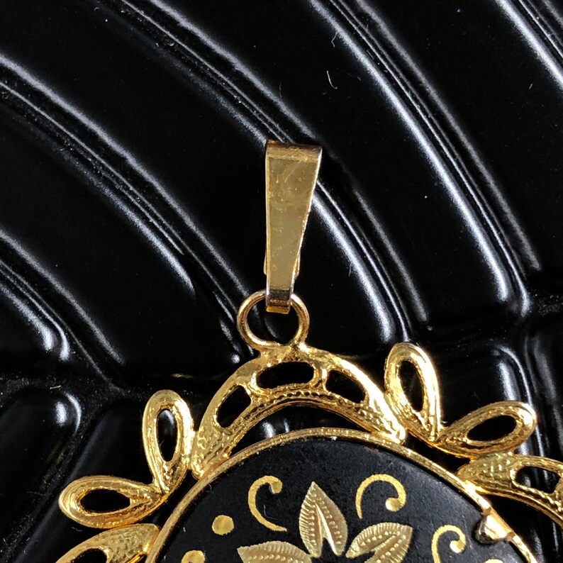 May include: A gold pendant with a black enamel center featuring a floral design. The pendant has a detailed, ornate frame and a loop for attaching a chain.