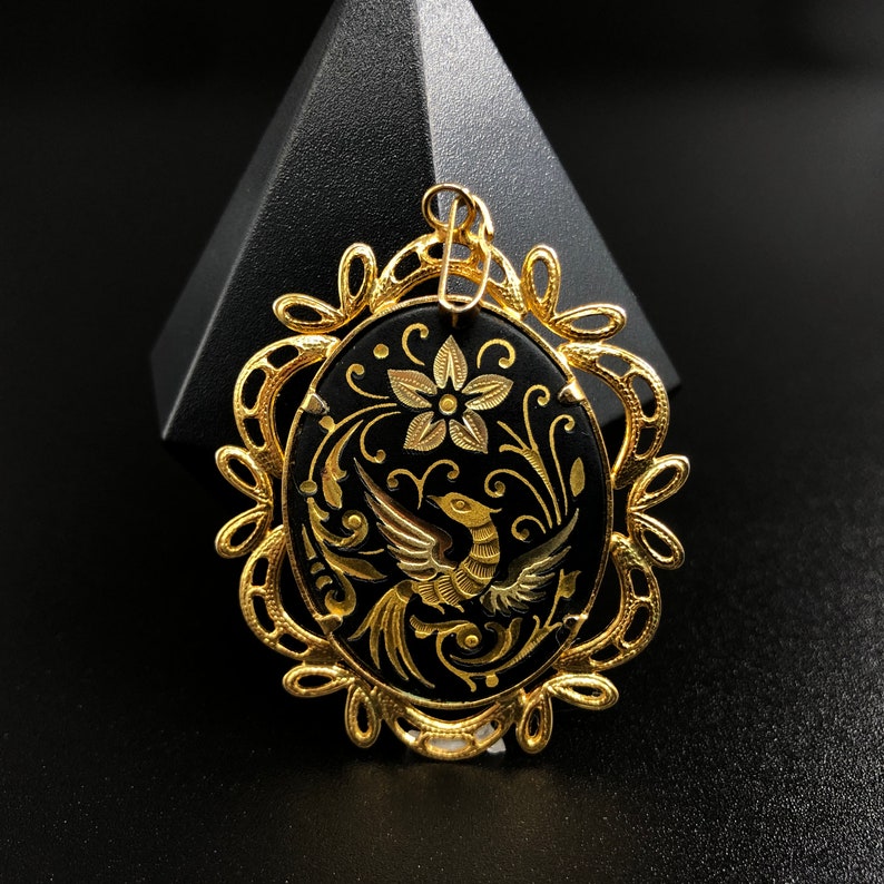May include: Gold pendant with a black enamel oval featuring a detailed design of a bird with floral accents.