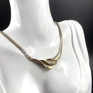 Pode incluir: Um colar de tom prateado com um design de corrente plana trançada que transita para um elemento central de metal liso e curvo com uma aparência dobrada em forma de onda. A peça repousa sobre um busto branco, exibindo o seu acabamento metálico.