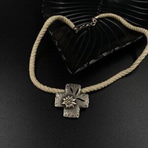Puede incluir: Un collar de plata con una cadena de cuerda y un colgante de cruz de plata con una flor de edelweiss dorada.