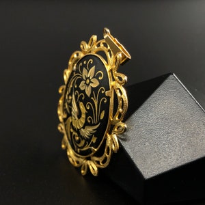 May include: A gold-tone pendant with a black oval cameo featuring a detailed floral design and a bird. The cameo is surrounded by a decorative gold frame.