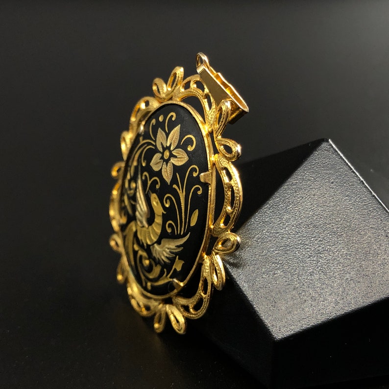 May include: A gold-tone pendant with a black oval cameo featuring a detailed floral design and a bird. The cameo is surrounded by a decorative gold frame.