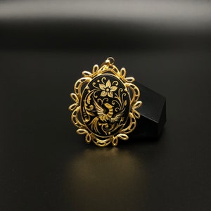 May include: A gold pendant with a black enamel oval featuring a stylized bird and floral design.