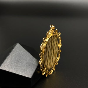 May include: Gold-toned oval-shaped pendant with a decorative filigree frame. The pendant has a smooth, textured surface.