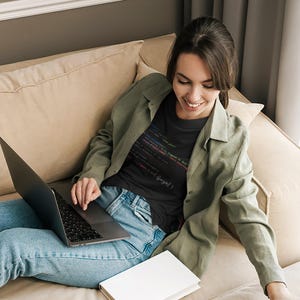 May include: A woman wearing a black t-shirt with colorful code text, light blue jeans, and a green jacket, using a laptop. A white book is on the couch. The woman is sitting on a beige couch.