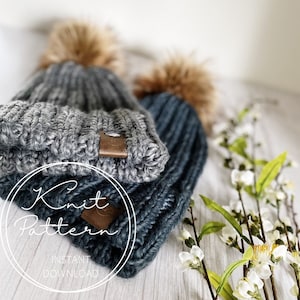 May include: Two knitted hats with faux fur pom poms. The hats are gray and blue with a brown leather button on the side. The hats are on a white wooden surface with white flowers.
