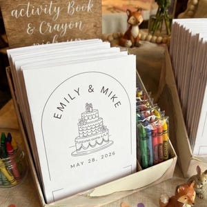 May include: A stack of white wedding activity books with a black line drawing of a three-tiered cake, the names Emily & Mike, and the date May 28, 2026. Each book is paired with a small clear plastic sleeve containing a set of colorful wax crayons.